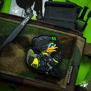 May include: A camouflage tactical pouch with a cartoon duck patch wearing sunglasses and a helmet. The patch has the text "zfd". Black Oakley sunglasses and a black magazine holder are also visible.