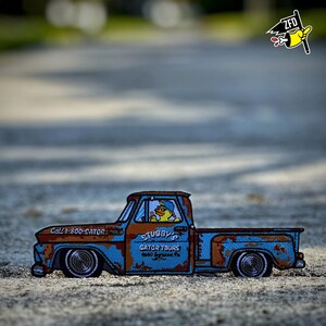 May include: An embroidered patch shaped like a rusted blue pickup truck with brown weathering. The truck features the text Call 1-800-GATOR and STUBBY'S GATOR TOURS. A yellow duck wearing a blue cap is visible in the driver's seat of the vehicle.