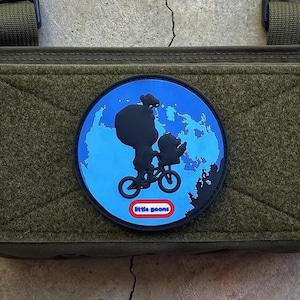 May include: A green tactical patch with a blue and white patch featuring a silhouette of a person riding a bicycle with the text "Little Goons" in red. The patch is attached to a green tactical vest with black straps and buckles.