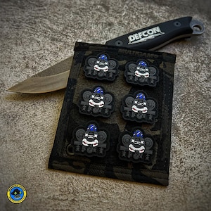 May include: Six black and white patches with a cartoon airplane design. The patches have blue accents and a skull and crossbones design. The patches are attached to a black and green camouflage fabric.