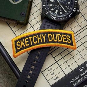 May include: A rectangular embroidered patch with a black background and a thick yellow border. The text SKETCHY DUDES is stitched in yellow capital letters across the center. The patch is positioned over a black watch strap and a watch face.