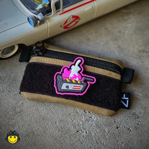 May include: A pink and white embroidered patch featuring a ghost trap with a ghost inside. The patch is attached to a brown and black zippered pouch with a black strap.