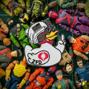 May include: A white embroidered patch of a duck with a red heart and the text "ZFD". The patch is surrounded by various toy action figures, including soldiers, robots, and a duck.