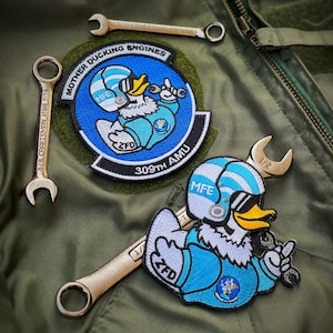 May include: Two embroidered patches featuring a cartoon duck wearing a helmet and holding a wrench. The patches are blue, white, and yellow. The text on the patches reads "Mother Ducking Engines" and "309th AMU".