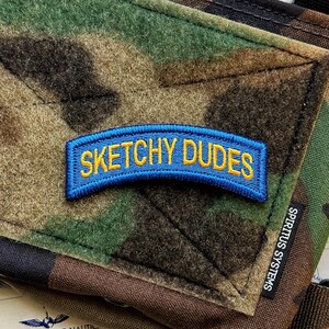 May include: A curved, blue embroidered patch with yellow text reading SKETCHY DUDES, attached to a camouflage fabric surface. A small black fabric tag with the white text SPIRITUS SYSTEMS is visible on the right side of the camouflage material.