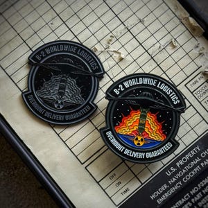 May include: Two circular black and gray metal pins. Each pin features the text "B-2 WORLDWIDE LOGISTICS" and "OVERNIGHT DELIVERY GUARANTEED". One pin depicts a bomb with smoke, the other a bomb with flames.