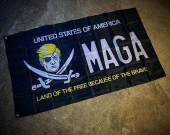May include: A black flag with a white and yellow design. The flag features a cartoon image of a person with a yellow hair and a black eyepatch. The text "UNITED STATES OF AMERICA" is at the top of the flag. The text "MAGA" is in large white letters in the center of the flag. The text "LAND OF THE FREE BECAUSE OF THE BRAVE" is at the bottom of the flag.