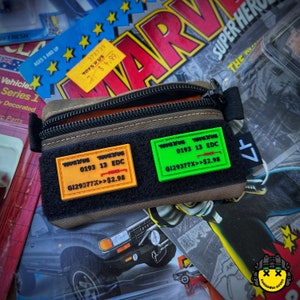 May include: A brown leather pouch with two orange and green rectangular patches. The patches have white text that reads "TOYS'R'US 0193 13 EDC G129377X>>$2.98".