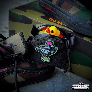 May include: A black zippered pouch with a black and white skeleton bird patch and a yellow rubber ducky inside. The pouch is attached to a pair of black and camouflage sneakers. The text "DIRTY BIRDS" is visible on the bottom right corner of the image.