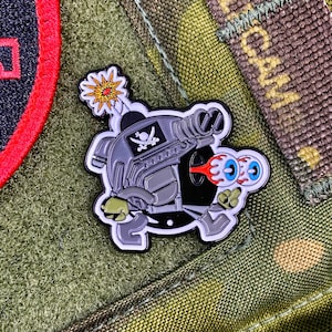 May include: A black and gray enamel pin featuring a cartoon character wearing a helmet with a skull and crossbones design. The character has one eye with a flame and the other eye is a cartoon eyeball. The pin is attached to a green and brown camouflage fabric.
