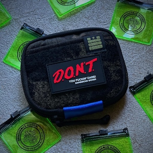 May include: A black rectangular zippered pouch featuring a rectangular patch with the red text D.O.N.T. and white text YOU FUCKIN' DARE! DANGEROUS GOODS. The pouch has a blue tab and is surrounded by several small, transparent neon green square pouches.