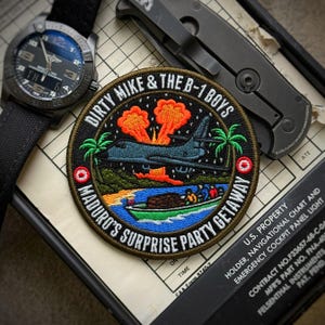 May include: A black watch with a black canvas strap, a patch with the text "DIRTY MIKE & THE B-1 BOYS MADURO'S SURPRISE PARTY GETAWAY", and a black folding knife are displayed on a map. The patch features a bomber plane, palm trees, and explosions.