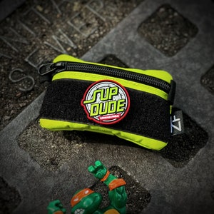 May include: A bright green and black zippered pouch with a black velcro strap. The pouch has a red and black circular patch with the words "Sup Dude" and "Cowabunga!" printed on it.