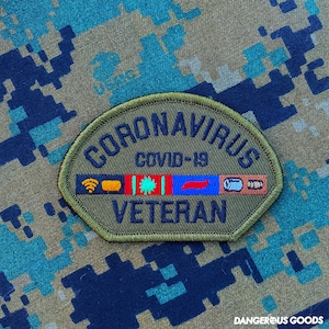 May include: An olive green embroidered patch with a curved top and flat bottom. The text reads CORONAVIRUS COVID-19 VETERAN in dark blue. A central horizontal strip features icons including a yellow wifi symbol, a green virus, and two pills.