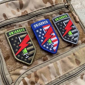 May include: Three embroidered patches with the word "UNJABBED" at the top. Each patch features a lightning bolt design in red, with stars and stripes or other graphic elements. The patches are in shades of black, blue, and gray.