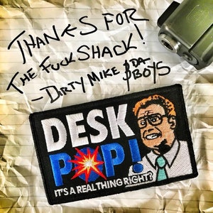 May include: A rectangular embroidered patch with a black background featuring a cartoon character with orange hair, glasses, and a teal tie. The text reads DESK POP! IT'S A REAL THING RIGHT? with a red and yellow starburst graphic between the letters.