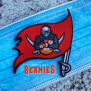 May include: A red and black embroidered patch featuring a pirate flag with a cartoon of Tom Brady wearing a Buccaneers jersey and holding a football. The text "Tampa Bay" and "Berrnies" is below the flag.