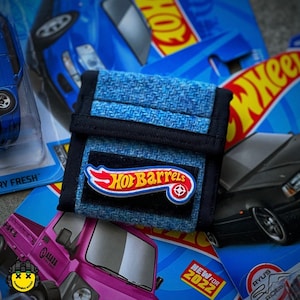 May include: A blue and black fabric wallet with a Hot Wheels logo that says "Hot Barrels" with a red, white, and blue design. The wallet is sitting on top of a Hot Wheels car toy packaging.