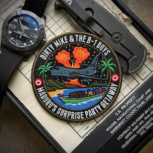 May include: Embroidered patch with the text "DIRTY MIKE & THE B-1 BOYS MADURO'S SURPRISE PARTY GETAWAY" featuring a B-1 bomber, palm trees, and a boat. A black watch and a folding knife are also visible.