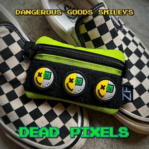 May include: A lime green zippered pouch with black trim and three black and yellow smiley face patches with pixelated designs. The pouch is sitting on top of a black and white checkered Vans shoe with the text "Dangerous Goods Smiley's" and "Dead Pixels" printed on the shoe.