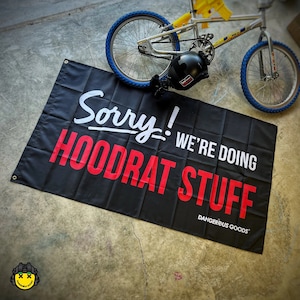 May include: A rectangular black banner featuring the white text Sorry! and the red text WE'RE DOING HOODRAT STUFF, with the white text DANGEROUS GOODS in the bottom right corner. The banner has metal grommets in the corners for hanging purposes.