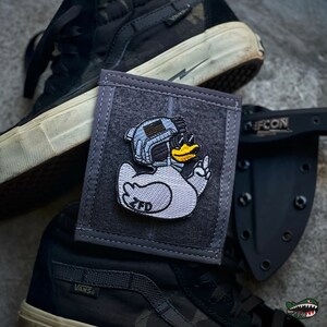 May include: A rectangular gray fabric patch featuring an embroidered white rubber duck wearing a tactical helmet with a small American flag and goggles. The duck has a yellow beak and the letters ZFD are stitched in black on its lower body.