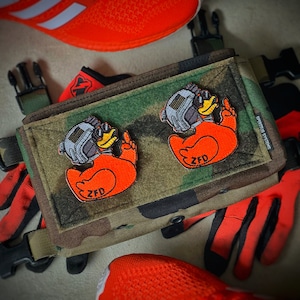 May include: A camouflage tactical pouch featuring two embroidered patches of orange ducks wearing tactical helmets and night vision goggles, each displaying the text ZFD. The pouch includes black plastic buckles and is surrounded by bright orange gear.