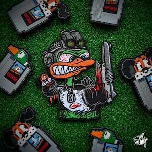 May include: A green, black, and white embroidered patch of a cartoon duck wearing a military helmet and holding a gun. The duck has red eyes and a menacing expression. The patch is surrounded by other patches with the text "DOG HUNT" and "DUCK HUNT" on them.