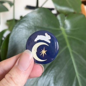 May include: A blue and gold enamel pin featuring a white rabbit jumping over a crescent moon with a star.