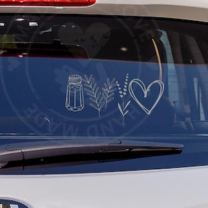 May include: White car rear window with a decal featuring a salt shaker, sprigs, and a heart. The decal is white and the car is parked outside with trees in the background.