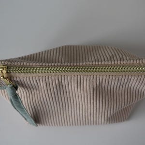 May include: A beige corduroy pouch with a gold-coloured zip and a small, light green fabric loop attached to the zip. The pouch has a rectangular shape with a curved top and is made of vertically ribbed corduroy fabric.