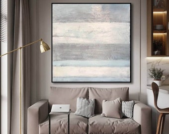 May include: A gray and white abstract painting with horizontal stripes hangs above a light gray couch with pillows. The painting is in a black frame and is on a wall in a modern living room.