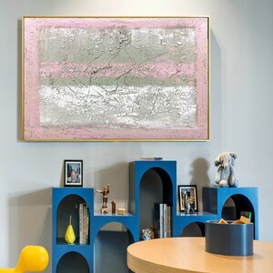 May include: Abstract painting with pink, white, and gray colors on a canvas. The painting is framed in gold and is hanging on a wall in a room with blue shelves and a wooden table.