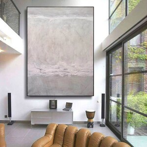 May include: A large abstract painting in shades of gray and white hangs on a white wall in a modern living room. The painting features a textured, layered effect, creating a sense of depth and movement. The painting is framed in a thin black frame.