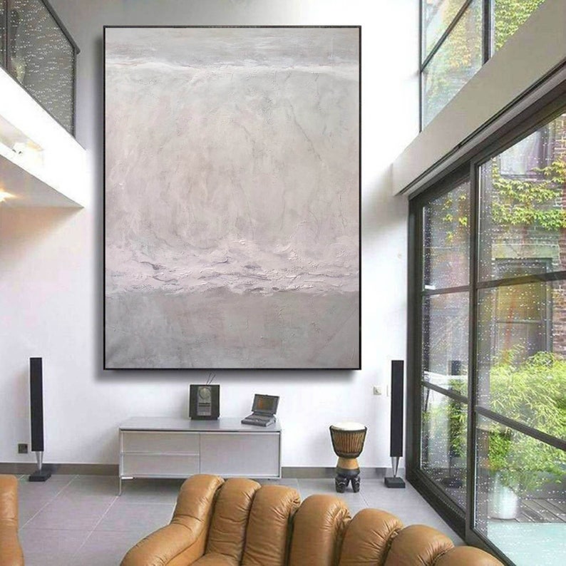 May include: A large abstract painting in shades of gray and white hangs on a white wall in a modern living room. The painting features a textured, layered effect, creating a sense of depth and movement. The painting is framed in a thin black frame.