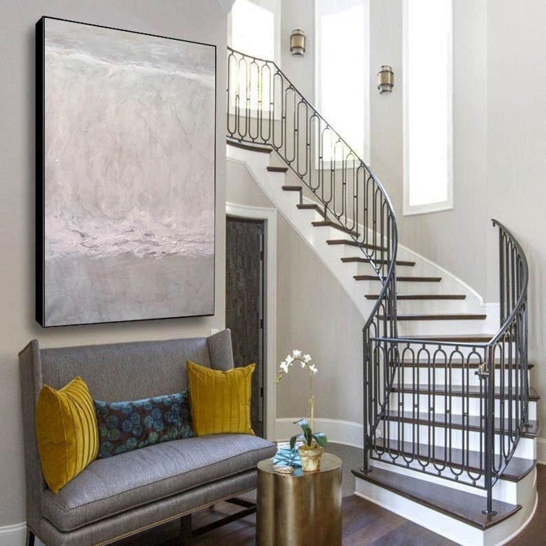 May include: A gray and white abstract painting hangs above a gray sofa with yellow and blue throw pillows. A wrought iron staircase with a white railing is in the background.