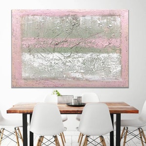 May include: Abstract painting with a textured, layered look. The painting features shades of pink, white, and gray. The painting is framed and hanging on a white wall above a wooden table with white chairs.
