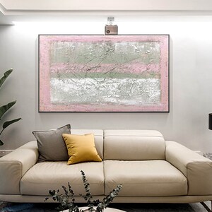 May include: Abstract painting with pink, white, and gray stripes on a canvas. The painting is framed in a black frame and is hanging on a white wall above a beige sofa.