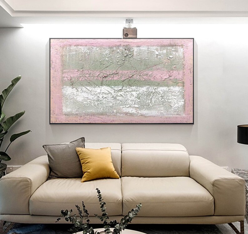 May include: Abstract painting with pink, white, and gray stripes on a canvas. The painting is framed in a black frame and is hanging on a white wall above a beige sofa.