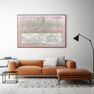 May include: Abstract painting with pink and gray stripes on a textured canvas. The painting is framed in black and hanging on a white wall above a brown leather sofa.