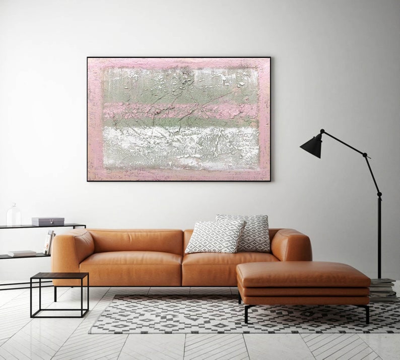 May include: Abstract painting with pink and gray stripes on a textured canvas. The painting is framed in black and hanging on a white wall above a brown leather sofa.
