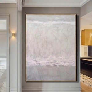 May include: Abstract painting with a textured, layered effect in shades of white, gray, and light pink. The painting is framed in a thin, gold frame and is hanging on a wall in a hallway.