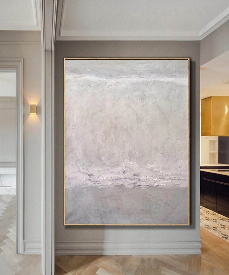 May include: Abstract painting with a textured, layered effect in shades of white, gray, and light pink. The painting is framed in a thin, gold frame and is hanging on a wall in a hallway.