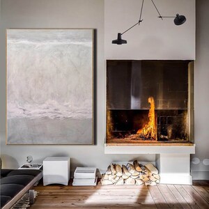 May include: A large abstract painting in shades of gray and white hangs on a wall in a modern living room. The painting depicts a landscape with a horizon line and clouds. A fireplace with a roaring fire is visible in the background.