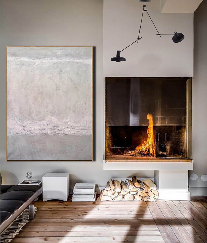 May include: A large abstract painting in shades of gray and white hangs on a wall in a modern living room. The painting depicts a landscape with a horizon line and clouds. A fireplace with a roaring fire is visible in the background.