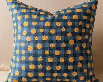 May include: A square throw pillow featuring a dark blue and light blue buffalo plaid pattern. The fabric is decorated with a repeating print of small, orange pumpkins distributed evenly across the entire surface of the pillow cover.
