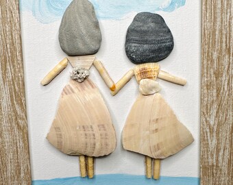 May include: A framed wall art piece featuring two figures crafted from smooth gray and dark charcoal stones, with bodies made of light-colored seashells. The figures hold hands against a painted blue sky and sea background with a small yellow heart.