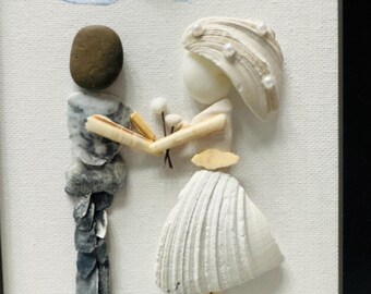 May include: A framed wall art piece featuring two figures crafted from seashells and stones. One figure is made of grey stones, the other of white shells with pearl accents. A painted blue sky with a shell heart and a blue base with a starfish.
