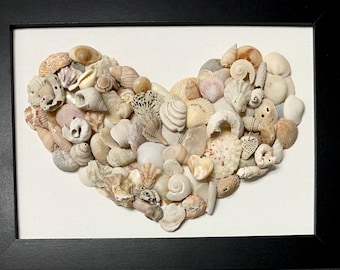 May include: A framed wall art piece featuring a collection of various seashells arranged in the shape of a heart. The shells are in shades of white, cream, beige, and tan, set against a white background within a simple black rectangular frame.