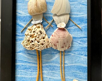May include: A framed wall art piece featuring two figures crafted from seashells. The figures have shell heads, porous coral-like skirts, and pink-tinted fan shell skirts, with thin yellow stems for legs, set against a blue wavy background.
